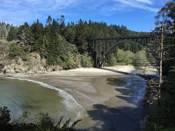 a creek passes under a bridge connecting steep, wooded banks, and meets the ocean at a sandy beach